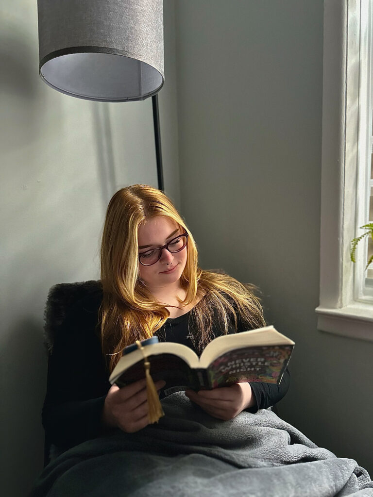 A woman sits reading a book next to a window. A lamp is above her, a blanket is over her lap.
